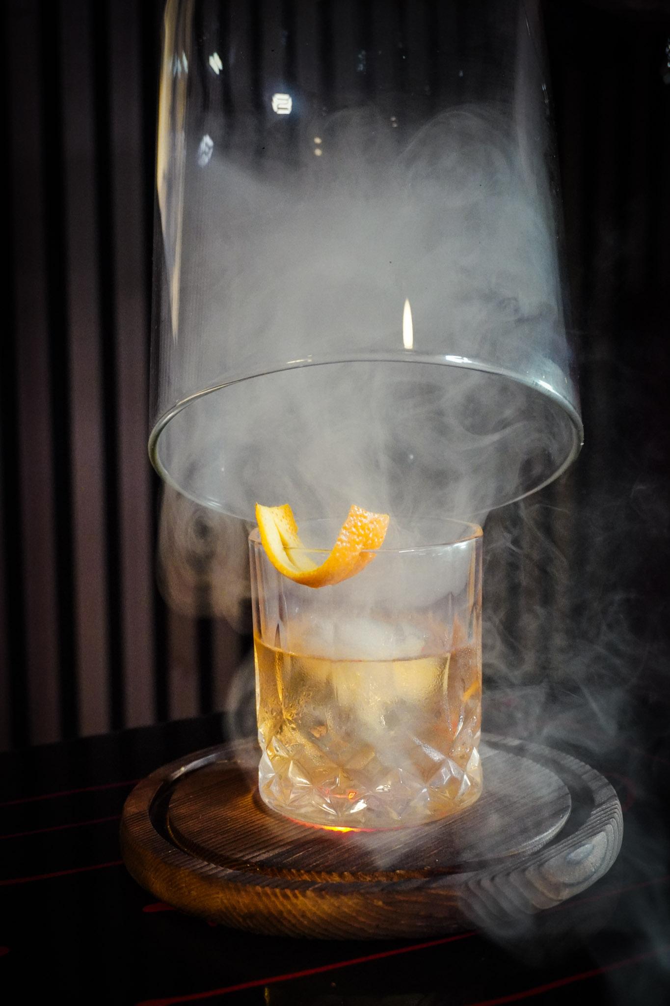 A smoky cocktail under a glass cloche on a wooden board.