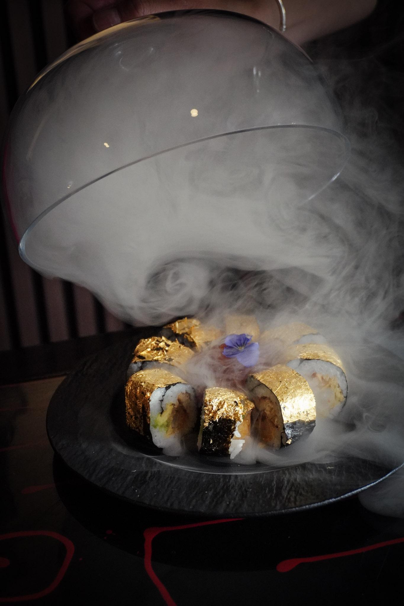 A gold leaf sushi roll revealed from under a glass dome with theatrical smoke.